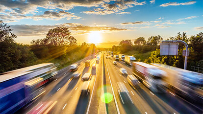 Auf einer Autobahn fahren Autos. Im Hintergrund sind Bäume und ein Sonnenuntergang zu sehen.