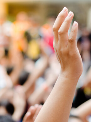 Eine Hand wird in die Höhe gehoben. Im Hintergrund sind weitere Menschen zu sehen, die ihre Hand heben.